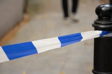 Fragment biało-niebieskiej taśmy policyjnej zawieszonej na czarnym słupku na chodniku, w tle nogi przechodnia.